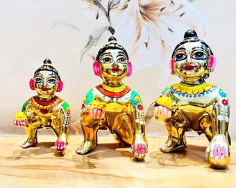 Può includere: Tre statuette dorate di una divinità, ciascuna in posa accovacciata. Hanno capelli neri, accenti rosa alle orecchie e dettagli di abbigliamento colorati. Le statuette sono su una superficie di legno, con uno sfondo floreale.