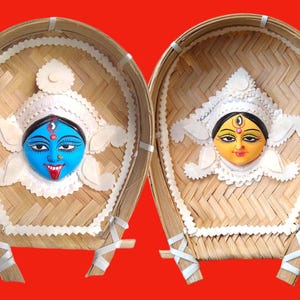 May include: Two decorative round woven bamboo trays with white trim, each featuring a painted face of a Hindu goddess. One goddess has blue skin and a red tongue, while the other has yellow skin and a red bindi.