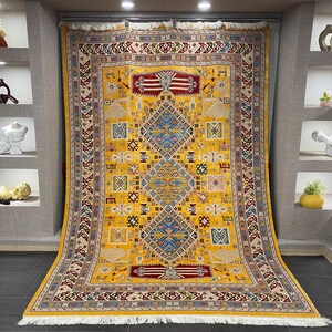 K&ouml;nnte beinhalten: Ein gro&szlig;er, rechteckiger gelber Teppich mit einem komplizierten geometrischen Muster. Der Teppich hat ein zentrales Rautenmuster in Blau und Rot, umgeben von kleineren Quadraten und Bord&uuml;ren in verschiedenen Farben. Der Teppich hat einen Fransenrand.