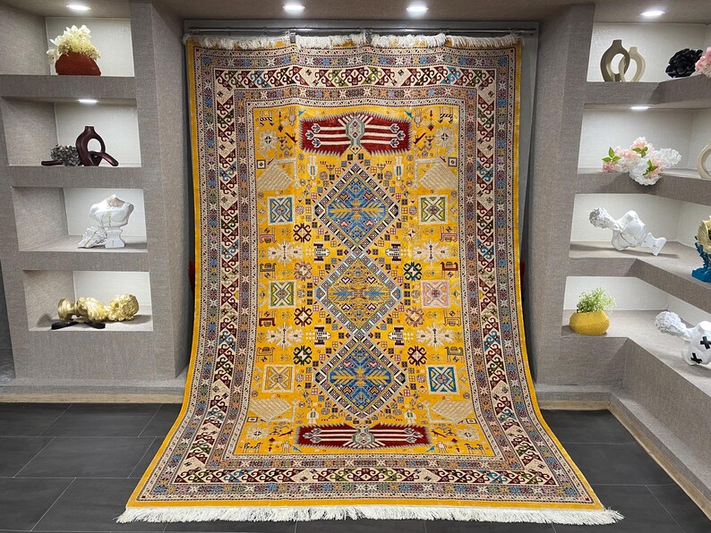 K&ouml;nnte beinhalten: Ein gro&szlig;er, rechteckiger gelber Teppich mit einem komplizierten geometrischen Muster. Der Teppich hat ein zentrales Rautenmuster in Blau und Rot, umgeben von kleineren Quadraten und Bord&uuml;ren in verschiedenen Farben. Der Teppich hat einen Fransenrand.