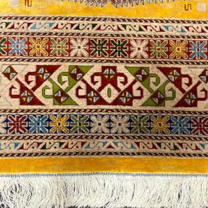 K&ouml;nnte beinhalten: Nahaufnahme eines gelb-cremefarbenen Teppichs mit komplizierten geometrischen Mustern. Der Teppich weist ein sich wiederholendes Design aus floralen und geometrischen Formen in Rot, Gr&uuml;n, Blau und Gelb auf. Der Teppich hat einen Fransenrand.