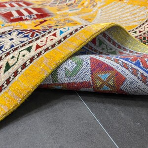 K&ouml;nnte beinhalten: Nahaufnahme eines aufgerollten, aufwendig gemusterten Teppichs. Der Teppich hat einen gelben Hintergrund mit roten, blauen und wei&szlig;en geometrischen Mustern. Ein leuchtend gelber Rand mit metallischem Glanz umrahmt die Kante und hebt die detaillierte Handwerkskunst hervor.