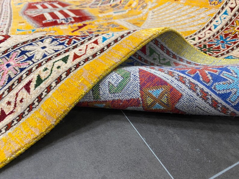 K&ouml;nnte beinhalten: Nahaufnahme eines aufgerollten, aufwendig gemusterten Teppichs. Der Teppich hat einen gelben Hintergrund mit roten, blauen und wei&szlig;en geometrischen Mustern. Ein leuchtend gelber Rand mit metallischem Glanz umrahmt die Kante und hebt die detaillierte Handwerkskunst hervor.