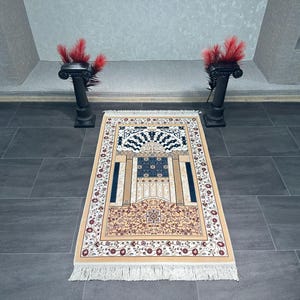 May include: A rectangular prayer rug with a beige border and a central design featuring a blue and beige archway. The rug has a floral pattern and a white fringe. Two black decorative pillars with red feathers are on either side.