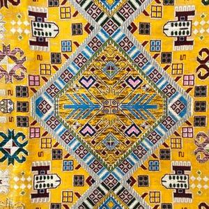 K&ouml;nnte beinhalten: Nahaufnahme eines gelben Teppichs mit einem komplizierten geometrischen Muster. Das Design zeigt Rauten, Quadrate und stilisierte Blumenmotive in Blau-, Rosa-, Braun- und Gr&uuml;nt&ouml;nen. Der Teppich hat ein detailliertes, strukturiertes Aussehen.