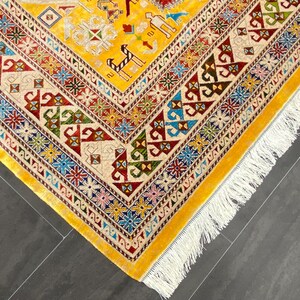 K&ouml;nnte beinhalten: Nahaufnahme eines gelben, aufwendig gemusterten Teppichs mit cremefarbenem Rand und wei&szlig;em Fransen. Der Teppich weist geometrische Designs in Rot, Blau, Gr&uuml;n und Braun auf. Die Ecke des Teppichs ist sichtbar und zeigt die detaillierte Handwerkskunst.