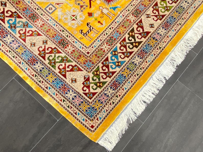 K&ouml;nnte beinhalten: Nahaufnahme eines gelben, aufwendig gemusterten Teppichs mit cremefarbenem Rand und wei&szlig;em Fransen. Der Teppich weist geometrische Designs in Rot, Blau, Gr&uuml;n und Braun auf. Die Ecke des Teppichs ist sichtbar und zeigt die detaillierte Handwerkskunst.