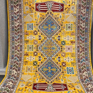 K&ouml;nnte beinhalten: Ein gro&szlig;er, rechteckiger gelber Teppich mit einem komplizierten geometrischen Muster. Der Teppich hat ein zentrales Rautenmuster in Blau und Wei&szlig;, umgeben von kleineren Quadraten und Formen in verschiedenen Farben. Der Rand hat ein detailliertes, sich wiederholendes Muster.