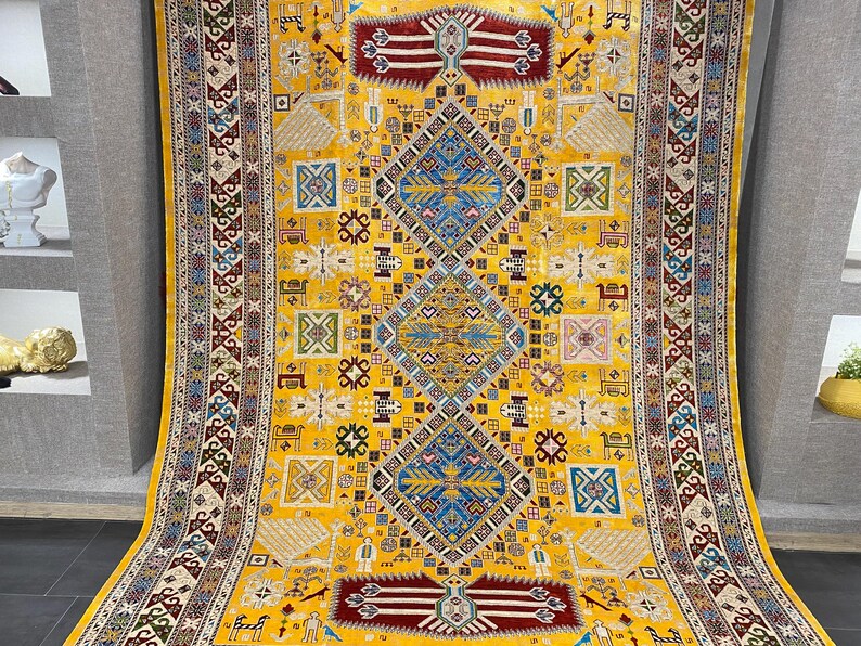 K&ouml;nnte beinhalten: Ein gro&szlig;er, rechteckiger gelber Teppich mit einem komplizierten geometrischen Muster. Der Teppich hat ein zentrales Rautenmuster in Blau und Wei&szlig;, umgeben von kleineren Quadraten und Formen in verschiedenen Farben. Der Rand hat ein detailliertes, sich wiederholendes Muster.