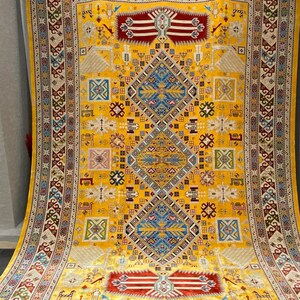 K&ouml;nnte beinhalten: Ein gro&szlig;er, verzierter Teppich mit einem leuchtend gelben Hintergrund. Der Teppich weist komplizierte geometrische Muster in Blau-, Rot- und Cremet&ouml;nen auf. Das Design umfasst Diamantenformen und Bord&uuml;ren mit detaillierten Motiven, wodurch ein reichhaltiges, strukturiertes Aussehen entsteht. Der Teppich ist wahrscheinlich aus Wolle oder einem &auml;hnlichen Material.