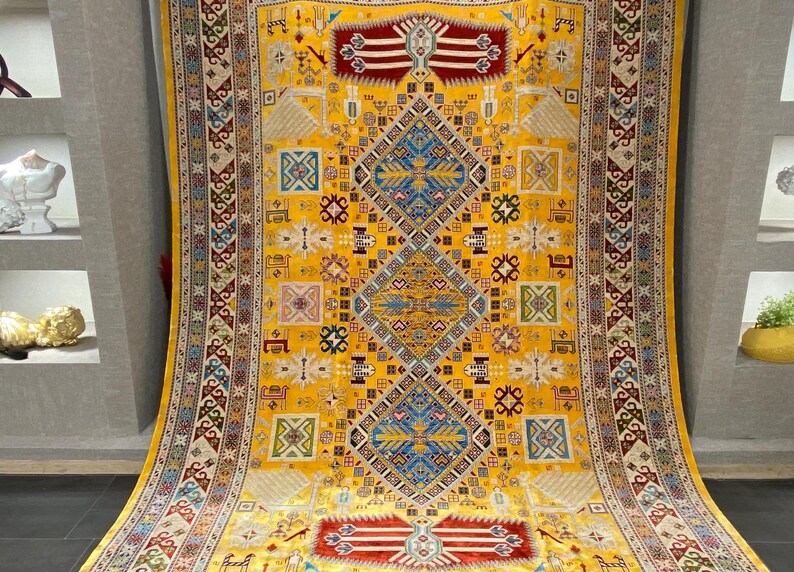 K&ouml;nnte beinhalten: Ein gro&szlig;er, verzierter Teppich mit einem leuchtend gelben Hintergrund. Der Teppich weist komplizierte geometrische Muster in Blau-, Rot- und Cremet&ouml;nen auf. Das Design umfasst Diamantenformen und Bord&uuml;ren mit detaillierten Motiven, wodurch ein reichhaltiges, strukturiertes Aussehen entsteht. Der Teppich ist wahrscheinlich aus Wolle oder einem &auml;hnlichen Material.