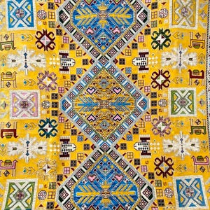 K&ouml;nnte beinhalten: Nahaufnahme eines gelben Teppichs mit einem aufwendigen geometrischen Muster. Das Design enth&auml;lt blaue, wei&szlig;e, rosa und gr&uuml;ne Elemente mit sich wiederholenden Rauten-, Quadrat- und Tiermotiven. Der Teppich hat einen Rand mit einem komplexen, farbenfrohen Design.