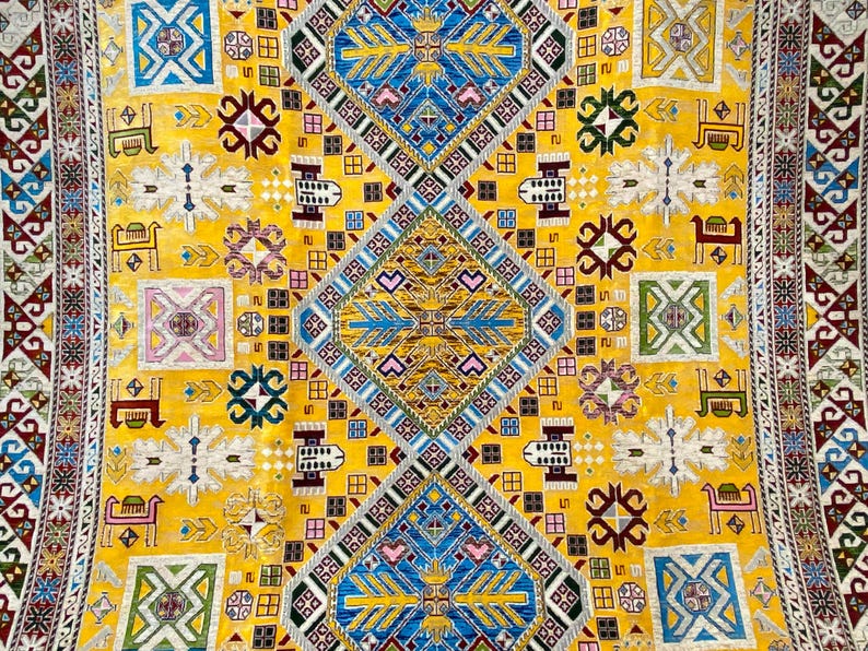 K&ouml;nnte beinhalten: Nahaufnahme eines gelben Teppichs mit einem aufwendigen geometrischen Muster. Das Design enth&auml;lt blaue, wei&szlig;e, rosa und gr&uuml;ne Elemente mit sich wiederholenden Rauten-, Quadrat- und Tiermotiven. Der Teppich hat einen Rand mit einem komplexen, farbenfrohen Design.
