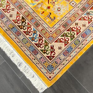 K&ouml;nnte beinhalten: Eine Ecke eines gelben Teppichs mit komplizierten geometrischen Mustern in Rot, Blau, Gr&uuml;n und Creme. Der Teppich hat einen cremefarbenen Rand mit farbenfrohen floralen Designs und einen Fransenrand. Das Design des Teppichs umfasst stilisierte Tierfiguren.
