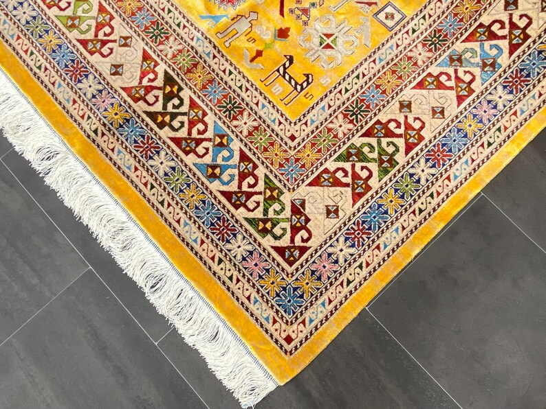 K&ouml;nnte beinhalten: Eine Ecke eines gelben Teppichs mit komplizierten geometrischen Mustern in Rot, Blau, Gr&uuml;n und Creme. Der Teppich hat einen cremefarbenen Rand mit farbenfrohen floralen Designs und einen Fransenrand. Das Design des Teppichs umfasst stilisierte Tierfiguren.