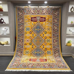 K&ouml;nnte beinhalten: Ein gro&szlig;er, rechteckiger gelber Teppich mit einem komplizierten geometrischen Muster. Der Teppich hat ein zentrales Rautenmuster in Blau und Rot, umgeben von kleineren Quadraten und Rechtecken in verschiedenen Farben. Der Teppich hat einen cremefarbenen Rand und eine Fransen am unteren Rand.