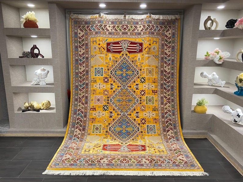 K&ouml;nnte beinhalten: Ein gro&szlig;er, rechteckiger gelber Teppich mit einem komplizierten geometrischen Muster. Der Teppich hat ein zentrales Rautenmuster in Blau und Rot, umgeben von kleineren Quadraten und Rechtecken in verschiedenen Farben. Der Teppich hat einen cremefarbenen Rand und eine Fransen am unteren Rand.