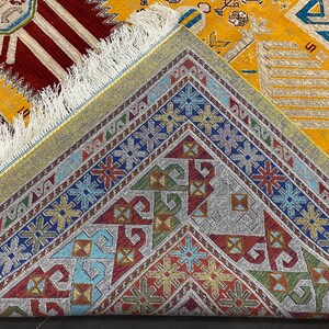 K&ouml;nnte beinhalten: Nahaufnahme eines gemusterten Teppichs mit gelbem Hintergrund. Der Teppich zeigt geometrische Muster in Rot, Blau, Gr&uuml;n und Gold mit einem Fransenrand. Die detaillierten Designs und leuchtenden Farben schaffen ein attraktives Textil.