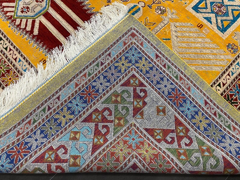 K&ouml;nnte beinhalten: Nahaufnahme eines gemusterten Teppichs mit gelbem Hintergrund. Der Teppich zeigt geometrische Muster in Rot, Blau, Gr&uuml;n und Gold mit einem Fransenrand. Die detaillierten Designs und leuchtenden Farben schaffen ein attraktives Textil.