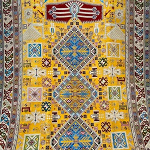 K&ouml;nnte beinhalten: Ein lebendiger gelber Teppich mit einem komplizierten geometrischen Design. Der Teppich hat ein zentrales Rautenmuster in Blau und Gold, umgeben von kleineren Quadraten und dekorativen Bord&uuml;ren in verschiedenen Farben. Das Design umfasst sich wiederholende Muster und Motive.