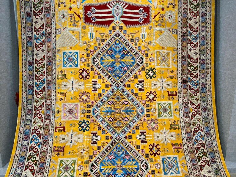 K&ouml;nnte beinhalten: Ein lebendiger gelber Teppich mit einem komplizierten geometrischen Design. Der Teppich hat ein zentrales Rautenmuster in Blau und Gold, umgeben von kleineren Quadraten und dekorativen Bord&uuml;ren in verschiedenen Farben. Das Design umfasst sich wiederholende Muster und Motive.