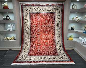 Könnte beinhalten: Ein großer, rechteckiger roter Teppich mit einem komplizierten Blumenmuster. Der Teppich hat einen cremefarbenen Rand mit geometrischen Designs und einen Fransenrand. Der Teppich wird vor einer neutralen Wand präsentiert und zeigt seine detaillierte Handwerkskunst.