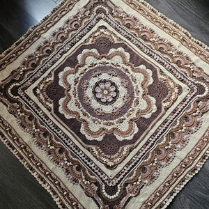 May include: A square crochet blanket featuring a complex mandala pattern in shades of cream, beige, and dark brown. The design includes concentric layers of intricate stitches, floral motifs, and textured bobbles, finished with a decorative border.
