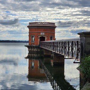 May include: A brick tower with a metal bridge leading to it, reflected in a still body of water under a cloudy sky.