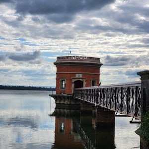 May include: A brick building with a bridge leading to it, standing on a body of water. The building has a flat roof and a small tower on top. The sky is cloudy and the water is reflecting the clouds.