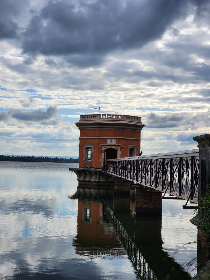 May include: A brick building with a bridge leading to it, standing on a body of water. The building has a flat roof and a small tower on top. The sky is cloudy and the water is reflecting the clouds.