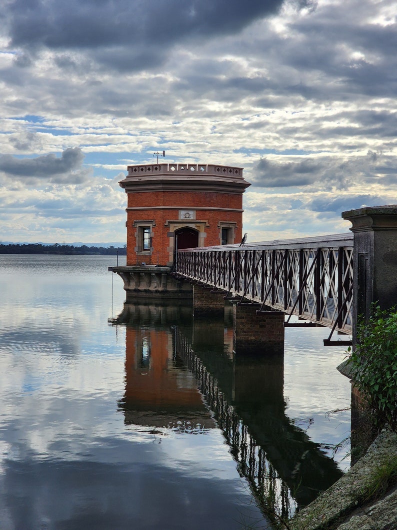 May include: A brick tower with a metal bridge leading to it, reflected in a still body of water under a cloudy sky.
