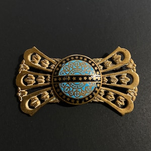 May include: A gold-toned metal buckle with a blue and black enamel design. The design features a circle with stars and a geometric pattern inside.