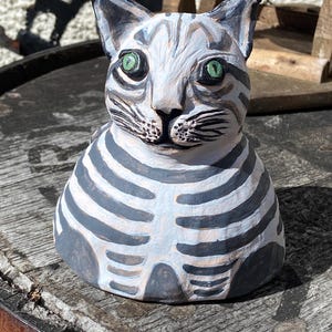 Może przedstawiać: Ręcznie malowana ceramiczna figurka kota z białym i szarym wzorem w paski. Kot ma zielone oczy i szczegółową twarz. Figurka to przedmiot dekoracyjny, prawdopodobnie do dekoracji domu.