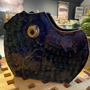 May include: A ceramic fish-shaped vase with a deep blue glaze. The vase has a prominent eye with a yellow iris and a textured surface. The design includes a fin and a mouth opening, suitable for holding flowers or other decorative items.