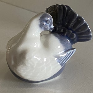 May include: A white porcelain figurine of a dove with a blue tail and wings. The dove is perched on its tail feathers and has a small beak.