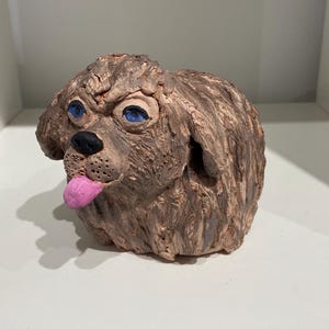 May include: A hand-sculpted clay figurine of a brown dog with a textured, shaggy coat. The dog has bright blue eyes, a black nose, and a pink tongue sticking out of its mouth. The sculpture features visible finger marks and a rough, organic finish.