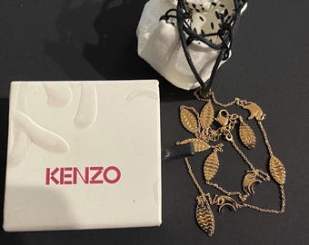 Könnte beinhalten: Eine goldene Halskette mit blattförmigen Anhängern und einem kleinen Elefantenanhänger. Die Halskette befindet sich in einer weißen Schachtel mit dem aufgedruckten Wort "KENZO".
