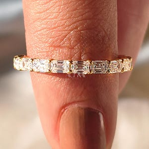 May include: A gold band ring set with a series of rectangular-cut, clear gemstones. The ring is held between two fingers, highlighting the ring's design and sparkle. The ring is a piece of jewellery.