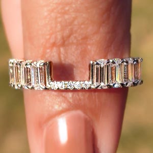 May include: A silver ring with a distinctive design. It features a row of rectangular-cut gemstones on the top and sides, with smaller stones in the centre. The ring is held by a finger, highlighting its elegant and modern style.
