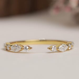 Puede incluir: Un anillo abierto de color dorado con una serie de pequeñas piedras preciosas transparentes. El anillo presenta un diseño único con un espacio abierto en el centro y piedras preciosas engastadas a lo largo de la banda. Las piedras preciosas incluyen tallas marquesa y redondas.