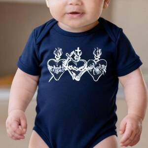 May include: A navy blue baby onesie with a white graphic of three hearts with a cross and flames. The baby is standing on a gray rug. The onesie has short sleeves and a round neckline.
