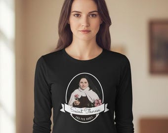 May include: A black long-sleeve shirt featuring a central oval graphic of Saint Therese in a habit holding roses. Below the oval, a white banner displays the text Saint Therese and the Latin phrase Ora Pro Nobis in black lettering.