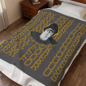 May include: A dark gray fleece blanket featuring a repeating gold text pattern that reads PRAY FOR US. In the center, there is a detailed illustration of Saint Charbel wearing a black habit and hood, with a white beard and a gold halo behind his head.