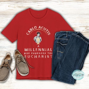 May include: A red t-shirt with the text "CARLO ACUTIS A MILLENNIAL WHO EMBRACED THE EUCHARIST" and an image of a person holding a chalice. The shirt is paired with blue jeans and gray shoes.