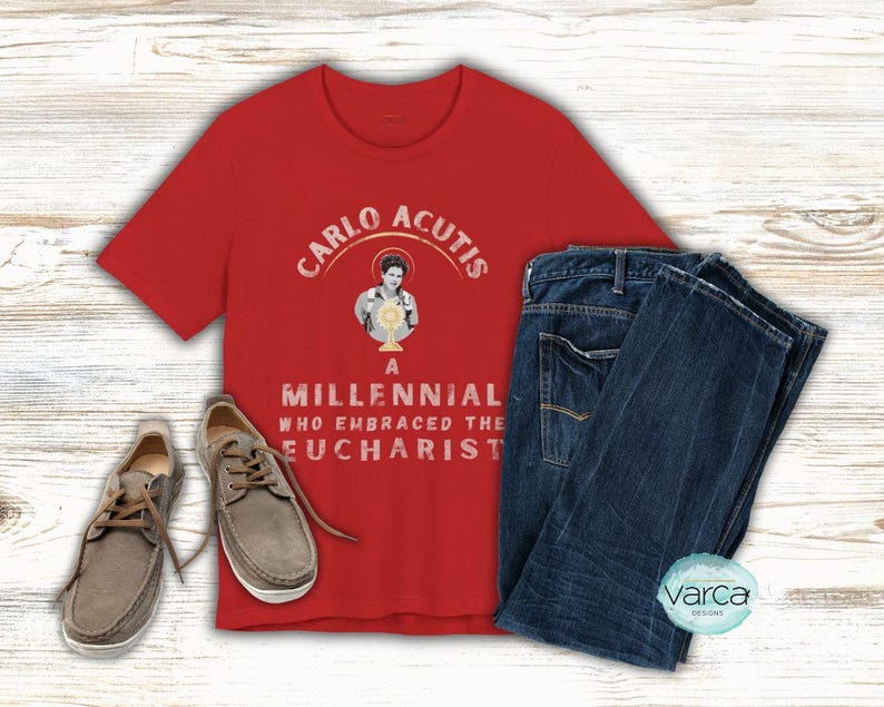 May include: A red t-shirt with the text "CARLO ACUTIS A MILLENNIAL WHO EMBRACED THE EUCHARIST" and an image of a person holding a chalice. The shirt is paired with blue jeans and gray shoes.