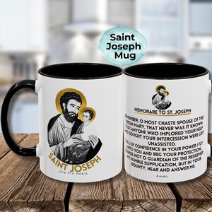 May include: White ceramic mug with a black handle and rim. The mug features a black and white image of Saint Joseph holding baby Jesus. The text "Saint Joseph Ora Pro Nobis" is printed below the image. The other side of the mug features a prayer to Saint Joseph.