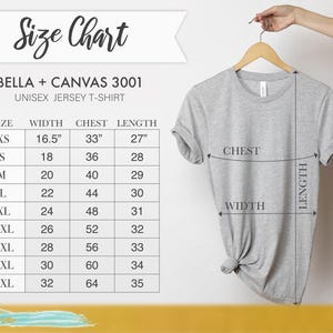 May include: A light gray Bella + Canvas 3001 unisex jersey t-shirt hanging on a wooden hanger. The image includes a size chart with measurements in inches for width, chest, and length, ranging from XS to 5XL.