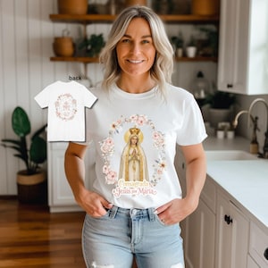 May include: White t-shirt featuring a religious illustration of a crowned figure within a floral wreath, with the text "Consagrada a Jesús por Maria." A smaller image of the back of the shirt is visible. The shirt is worn by a person.