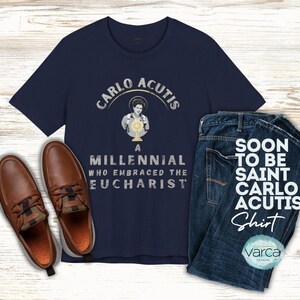 May include: Navy blue t-shirt with the text "CARLO ACUTIS" and an image of a person holding a chalice. The shirt is paired with blue jeans and brown leather shoes. The text "SOON TO BE SAINT CARLO ACUTIS Shirt" is also visible.
