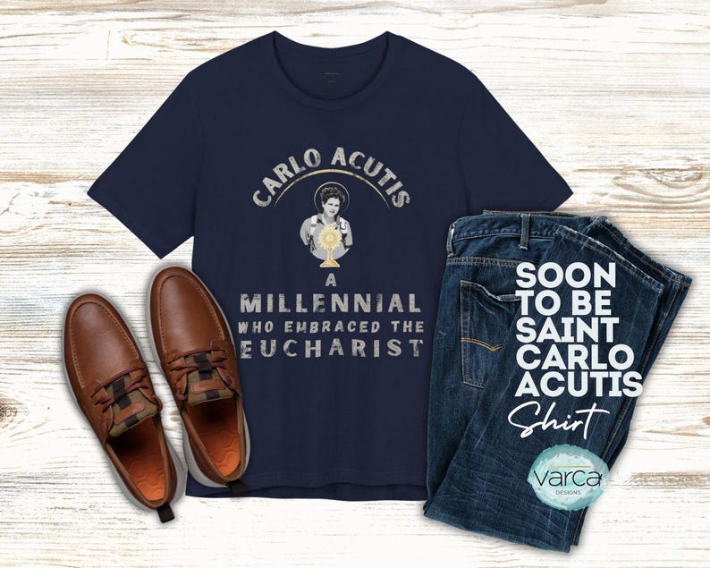 May include: Navy blue t-shirt with the text "CARLO ACUTIS" and an image of a person holding a chalice. The shirt is paired with blue jeans and brown leather shoes. The text "SOON TO BE SAINT CARLO ACUTIS Shirt" is also visible.